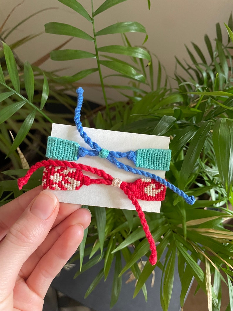 Peut inclure: Trois bracelets d'amiti&eacute; faits &agrave; la main dans des tons de bleu, turquoise et rouge. Les bracelets sont pr&eacute;sent&eacute;s sur une carte blanche sur fond de feuillage vert. Les bracelets sont faits de fils tiss&eacute;s et pr&eacute;sentent des motifs complexes.