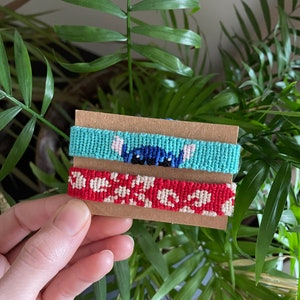 Peut inclure: Deux bracelets d'amiti&eacute; faits &agrave; la main sur une carte marron. L'un est bleu clair avec un motif de personnage de dessin anim&eacute;, l'autre rouge avec un motif floral blanc. Les bracelets sont faits de fil tiss&eacute;.