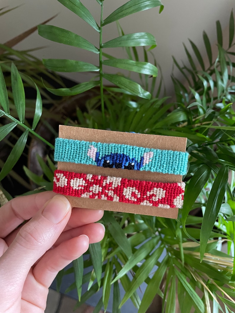 Peut inclure: Deux bracelets d'amiti&eacute; faits &agrave; la main sur une carte marron. L'un est bleu clair avec un motif de personnage de dessin anim&eacute;, l'autre rouge avec un motif floral blanc. Les bracelets sont faits de fil tiss&eacute;.