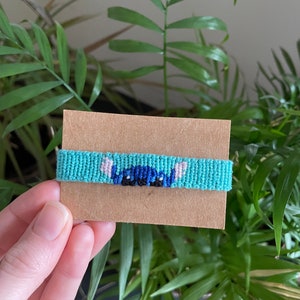 Peut inclure: Bracelet d'amiti&eacute; fait &agrave; la main, turquoise, avec un motif de personnage de dessin anim&eacute; bleu et noir. Le bracelet est pr&eacute;sent&eacute; sur une carte marron, avec des plantes vertes en arri&egrave;re-plan.