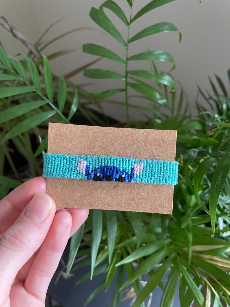 Peut inclure: Bracelet d'amiti&eacute; fait &agrave; la main, turquoise, avec un motif de personnage de dessin anim&eacute; bleu et noir. Le bracelet est pr&eacute;sent&eacute; sur une carte marron, avec des plantes vertes en arri&egrave;re-plan.