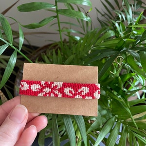 Peut inclure: Un bracelet en macram&eacute; rouge et blanc avec un motif floral est pr&eacute;sent&eacute; sur une carte marron. Le bracelet est tenu devant une plante verte luxuriante. Le motif en macram&eacute; pr&eacute;sente un motif r&eacute;p&eacute;titif de fleurs blanches sur fond rouge.