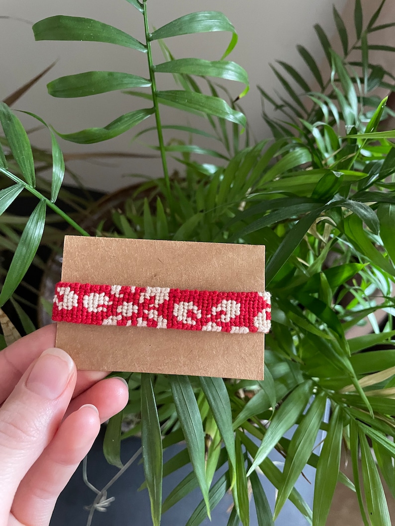 Peut inclure: Un bracelet en macram&eacute; rouge et blanc avec un motif floral est pr&eacute;sent&eacute; sur une carte marron. Le bracelet est tenu devant une plante verte luxuriante. Le motif en macram&eacute; pr&eacute;sente un motif r&eacute;p&eacute;titif de fleurs blanches sur fond rouge.
