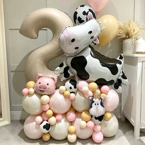 Könnte beinhalten: Eine Ballon-Arrangement mit einer großen Zahl zwei Ballon, einem Kuh-Ballon, einem Schwein-Ballon und einem kleineren Kuh-Ballon. Die Ballons sind von kleineren rosa, weißen und beigefarbenen Ballons umgeben.