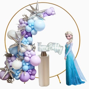 Puede incluir: Un arco de metal dorado decorado con una cascada de globos morados, azules y blancos. Un soporte para pastel blanco con un letrero que dice "Happy Birthday" est&aacute; frente al arco. Una figura de una ni&ntilde;a con un vestido azul se encuentra a la derecha del arco.