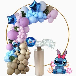 Puede incluir: Un arco de globos dorado con globos azules, morados y beige. El arco está decorado con hojas verdes y un personaje azul con un lazo rojo. Un soporte de pastel blanco con un letrero de "Feliz cumpleaños" está frente al arco.