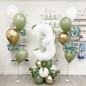 May include: A white number three balloon with the name Ollie written on it, surrounded by green, gold, and clear balloons with leaf patterns. The balloons are arranged in a cluster on a white surface.