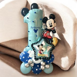 May include: A blue number one balloon with black Mickey Mouse ears and a Mickey Mouse character holding a star balloon. The balloon has white polka dots and the name "Anthony" written on it. The balloon is surrounded by smaller blue, white, and silver balloons.