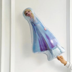 Puede incluir: Un globo de aluminio azul y morado con forma de Elsa de la pel&iacute;cula Frozen. Elsa lleva un vestido azul y morado con una capa blanca. Tiene el pelo largo y rubio y est&aacute; mirando hacia arriba.