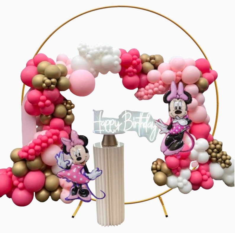 May include: A gold metal hoop arch decorated with pink, white, and gold balloons. Two Minnie Mouse figures stand in front of the arch, one holding a sign that says "Happy Birthday".