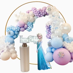 Peut inclure: Une arche en or décorée de ballons bleus, blancs et violets, de flocons de neige blancs et d'une pancarte indiquant "Joyeux anniversaire". Un costume d'Elsa du film La Reine des Neiges, bleu et blanc, est placé devant l'arche.