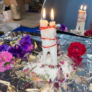 May include: A white wax candle shaped like two figures standing together, wrapped in red string and pierced with small metal pins. The candle has two lit wicks and is surrounded by scattered dried flower petals, herbs, and salt on foil.