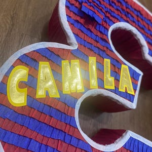 Puede incluir: Una pi&ntilde;ata grande, tridimensional con forma del n&uacute;mero 3, decorada con rayas alternas de papel crep&eacute; rojo y azul. El nombre "CAMILA" est&aacute; escrito en letras amarillas en la parte delantera. Los bordes est&aacute;n delineados en blanco.