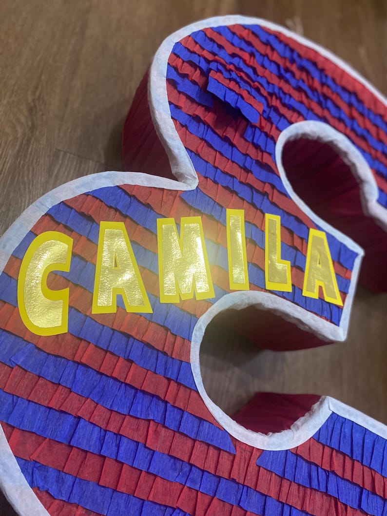 Puede incluir: Una pi&ntilde;ata grande, tridimensional con forma del n&uacute;mero 3, decorada con rayas alternas de papel crep&eacute; rojo y azul. El nombre "CAMILA" est&aacute; escrito en letras amarillas en la parte delantera. Los bordes est&aacute;n delineados en blanco.