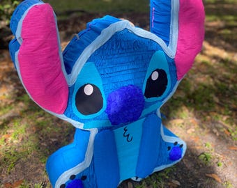 Op de afbeelding: Blauwe en roze Stitch-personage piñata. De piñata is gemaakt van crêpepapier en heeft een blauw lichaam, roze oren en witte ogen.