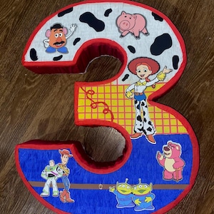 Puede incluir: Una pi&ntilde;ata grande con forma del n&uacute;mero 3, decorada con personajes de la pel&iacute;cula Toy Story. El n&uacute;mero est&aacute; delineado en rojo y relleno con papel azul, amarillo y blanco. Los personajes incluyen a Sr. Potato, Jessie y Buzz Lightyear.