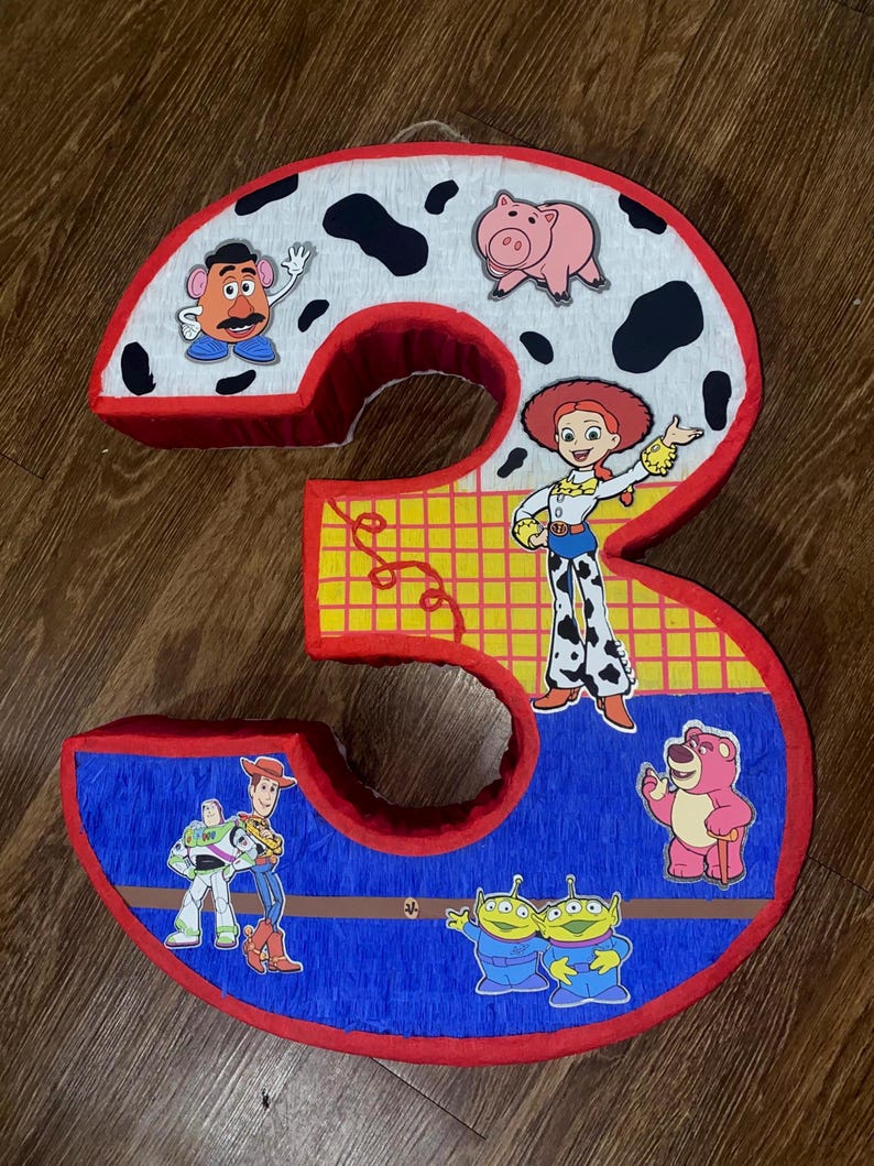 Puede incluir: Una pi&ntilde;ata grande con forma del n&uacute;mero 3, decorada con personajes de la pel&iacute;cula Toy Story. El n&uacute;mero est&aacute; delineado en rojo y relleno con papel azul, amarillo y blanco. Los personajes incluyen a Sr. Potato, Jessie y Buzz Lightyear.