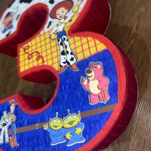 Puede incluir: Una pi&ntilde;ata grande y tridimensional con el n&uacute;mero "3", con personajes de la franquicia Toy Story. El n&uacute;mero es rojo, azul y amarillo, con pegatinas de personajes como Jessie, Lotso y los peque&ntilde;os hombrecillos verdes. La parte superior es blanca con manchas negras.