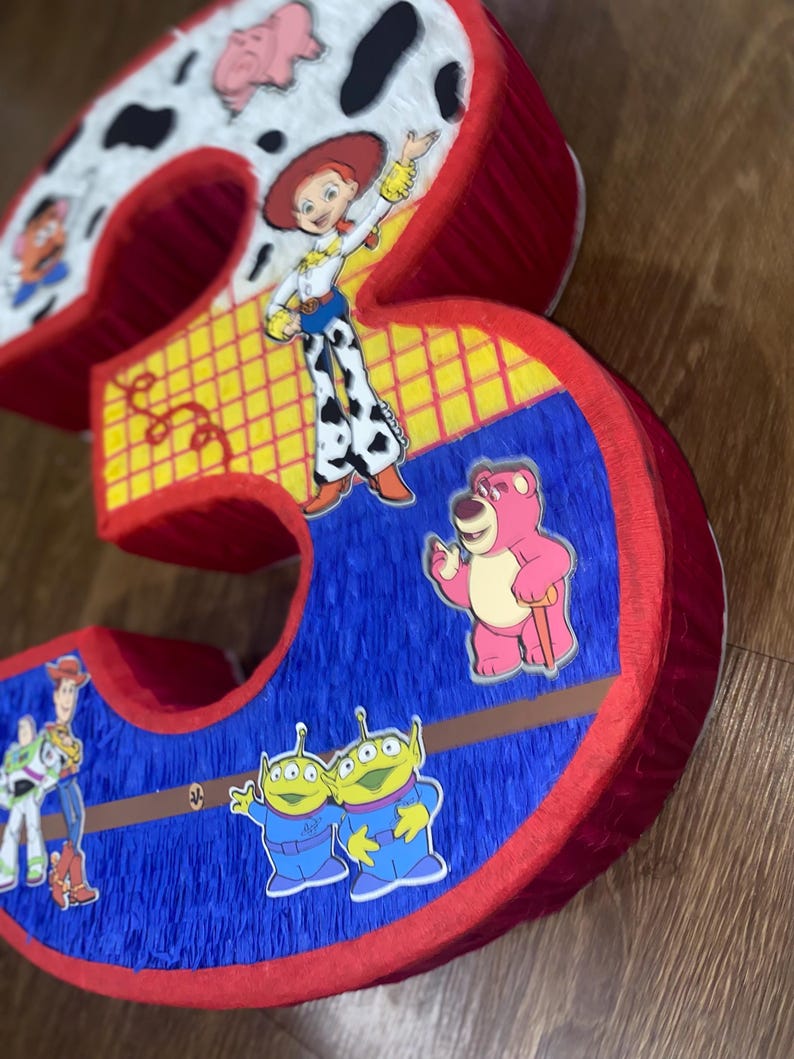 Puede incluir: Una pi&ntilde;ata grande y tridimensional con el n&uacute;mero "3", con personajes de la franquicia Toy Story. El n&uacute;mero es rojo, azul y amarillo, con pegatinas de personajes como Jessie, Lotso y los peque&ntilde;os hombrecillos verdes. La parte superior es blanca con manchas negras.
