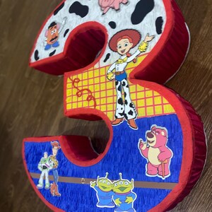 Puede incluir: Una pi&ntilde;ata con forma del n&uacute;mero tres con personajes de la pel&iacute;cula Toy Story. La pi&ntilde;ata es roja, blanca, azul y amarilla, con im&aacute;genes de Jessie, Buzz Lightyear y otros personajes.