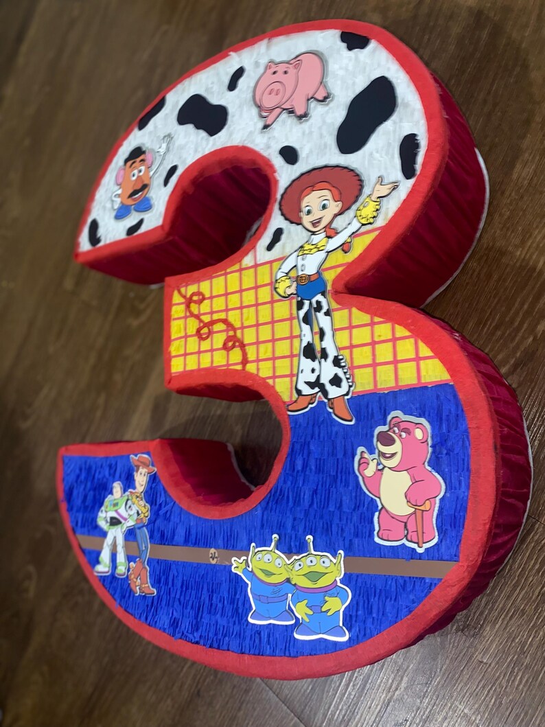 Puede incluir: Una pi&ntilde;ata con forma del n&uacute;mero tres con personajes de la pel&iacute;cula Toy Story. La pi&ntilde;ata es roja, blanca, azul y amarilla, con im&aacute;genes de Jessie, Buzz Lightyear y otros personajes.