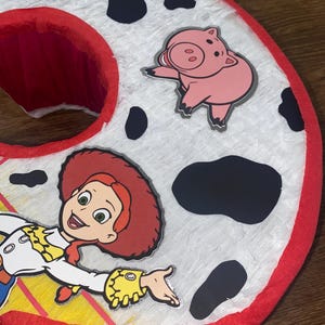 Puede incluir: Una pi&ntilde;ata grande, roja y blanca con el n&uacute;mero seis, con manchas negras y recortes de personajes de Toy Story. La pi&ntilde;ata presenta a Jessie la vaquera y un personaje de cerdo rosa. Los bordes est&aacute;n adornados con papel crep&eacute; rojo.