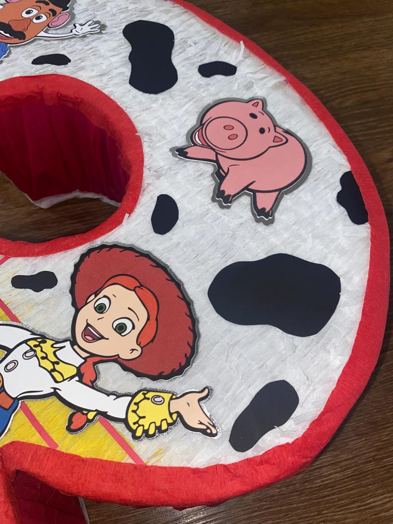 Puede incluir: Una pi&ntilde;ata grande, roja y blanca con el n&uacute;mero seis, con manchas negras y recortes de personajes de Toy Story. La pi&ntilde;ata presenta a Jessie la vaquera y un personaje de cerdo rosa. Los bordes est&aacute;n adornados con papel crep&eacute; rojo.