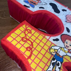 Puede incluir: Una pi&ntilde;ata roja y amarilla con forma del n&uacute;mero 2, decorada con personajes de la pel&iacute;cula Toy Story. La pi&ntilde;ata tiene un borde rojo y est&aacute; decorada con un estampado de vaca y el personaje Sr. Cara de Papa.