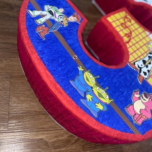 Puede incluir: Una pi&ntilde;ata grande con forma de letra 'C', con bordes rojos y una superficie azul texturizada. La pi&ntilde;ata est&aacute; decorada con im&aacute;genes de personajes de la pel&iacute;cula animada 'Toy Story', incluyendo a Buzz Lightyear, Woody y los marcianos.