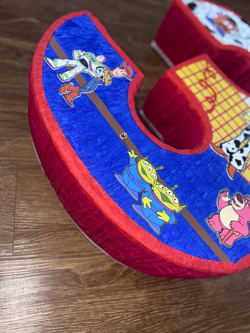 Puede incluir: Una pi&ntilde;ata grande con forma de letra 'C', con bordes rojos y una superficie azul texturizada. La pi&ntilde;ata est&aacute; decorada con im&aacute;genes de personajes de la pel&iacute;cula animada 'Toy Story', incluyendo a Buzz Lightyear, Woody y los marcianos.