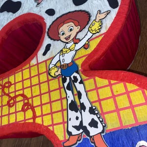 Puede incluir: Una pi&ntilde;ata hecha a mano con forma del n&uacute;mero nueve, con el personaje Jessie de Toy Story. La pi&ntilde;ata tiene una combinaci&oacute;n de colores rojo, amarillo y blanco con detalles de estampado de vaca y una ilustraci&oacute;n de cerdo rosa.