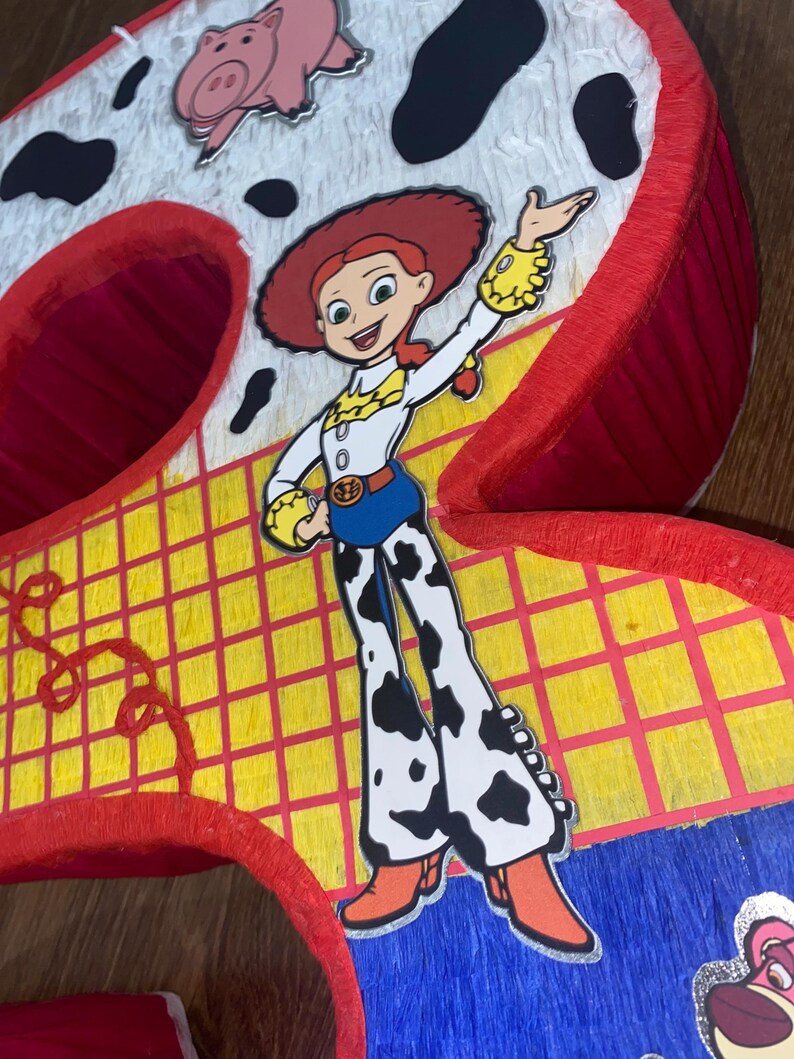 Puede incluir: Una pi&ntilde;ata hecha a mano con forma del n&uacute;mero nueve, con el personaje Jessie de Toy Story. La pi&ntilde;ata tiene una combinaci&oacute;n de colores rojo, amarillo y blanco con detalles de estampado de vaca y una ilustraci&oacute;n de cerdo rosa.