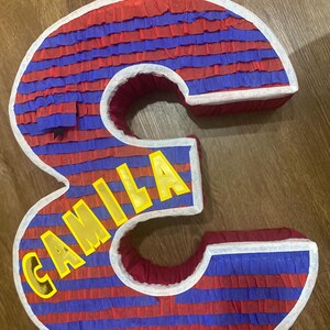 Puede incluir: Una pi&ntilde;ata grande con forma de n&uacute;mero 3 con rayas horizontales rojas y azules. La palabra "CAMILA" se muestra en letras amarillas. La pi&ntilde;ata est&aacute; bordeada con blanco y tiene un lazo para colgar.