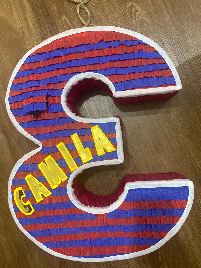 Puede incluir: Una pi&ntilde;ata grande con forma de n&uacute;mero 3 con rayas horizontales rojas y azules. La palabra "CAMILA" se muestra en letras amarillas. La pi&ntilde;ata est&aacute; bordeada con blanco y tiene un lazo para colgar.