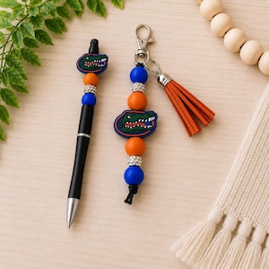 May include: A black ink pen and a matching keychain featuring orange and blue silicone beads, silver rhinestone spacers, and a rubber alligator mascot charm. The keychain includes a silver lobster clasp and a long orange faux leather tassel.