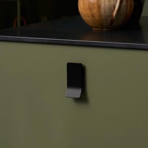 May include: An olive green kitchen cabinet with a black worktop. A small, round, wooden stool is in the lower left corner. On the worktop are a brown ceramic pitcher, a black ceramic jug, a bottle of liquor, and a small cup with utensils.