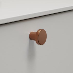 May include: A white cabinet drawer with a copper-coloured, round knob. The knob is centrally positioned on the drawer. The cabinet is likely part of a bathroom vanity or similar furniture.
