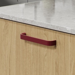 May include: A kitchen cabinet with a textured, claret-coloured handle. The cabinet is made of light-coloured wood and is set against a grey countertop. A stainless steel sink and tap are visible in the background.