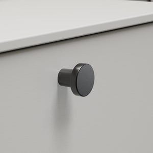 May include: A grey cabinet door with a black, circular knob. The cabinet is part of a bathroom vanity unit. The background includes a white countertop, a bottle of lotion, and a tube of cream.