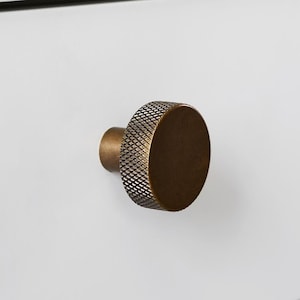 May include: A bronze-coloured drawer knob with a textured, circular design. The knob is mounted on a white drawer, creating a clean, modern look. The hardware adds a touch of elegance to the furniture. The knob is approximately 3 cm in diameter.