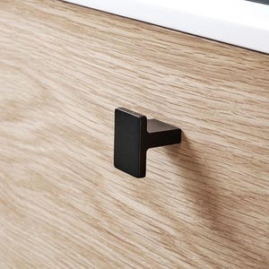 May include: A close-up of a kitchen cabinet with a light wood grain finish. The cabinet has a black, rectangular handle. A white countertop and a black hob are visible in the background.
