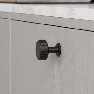 May include: A close-up shot of a kitchen cabinet door with a black, textured knob. The knob has a round, ridged design and a short stem. The cabinet is light grey, with a countertop and a black pot visible in the background.