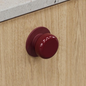 May include: A close-up of a red cabinet knob with a circular base and a rounded, cylindrical handle. The knob is mounted on a light-coloured wooden cabinet door. A light-coloured countertop is visible in the background.