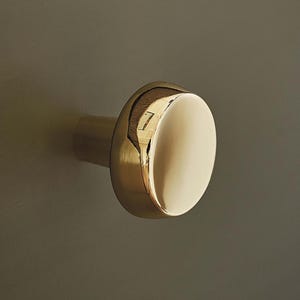 May include: A close-up of a gold cabinet knob with a rounded, flat face. The knob is mounted on a muted green surface. The knob's surface reflects light, creating highlights and shadows.