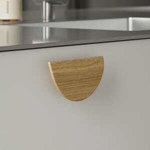 May include: A light-coloured, semi-circular wooden cabinet pull is mounted on a light grey cabinet door. The pull has a natural wood grain pattern. The kitchen setting includes a stainless steel sink and tap.