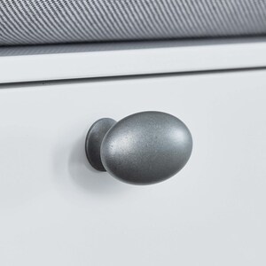 May include: A white cabinet drawer with a grey oval knob. A grey and white patterned cushion sits above. The metal knob has a matte finish. The cushion has a textured appearance. The cabinet is likely part of a larger piece of furniture.