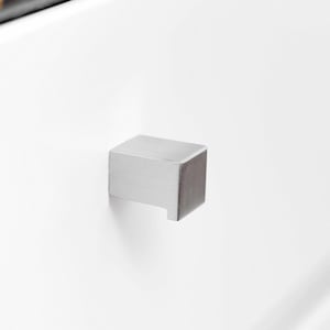 May include: A brushed silver cabinet pull with a square design. The pull is mounted on a white cabinet door. A dark countertop is visible in the background.