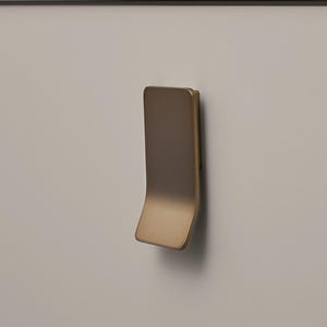 May include: A contemporary brass cabinet pull with a curved design, mounted on a light grey cabinet door. The handle's warm metallic finish contrasts with the neutral door. The curved shape offers a minimalist and stylish aesthetic, suitable for modern interiors.