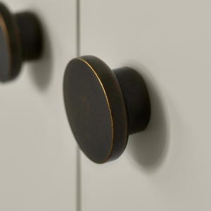 May include: Close-up of two dark bronze cabinet knobs with a gold-coloured edge. The knobs are round and mounted on a light grey surface. The knobs are a decorative hardware element.