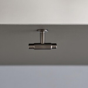 May include: A brushed metal cabinet pull with a T-shaped design. The horizontal bar has a ridged texture, and the pull is mounted on a grey surface. The background is a gradient of grey and white.
